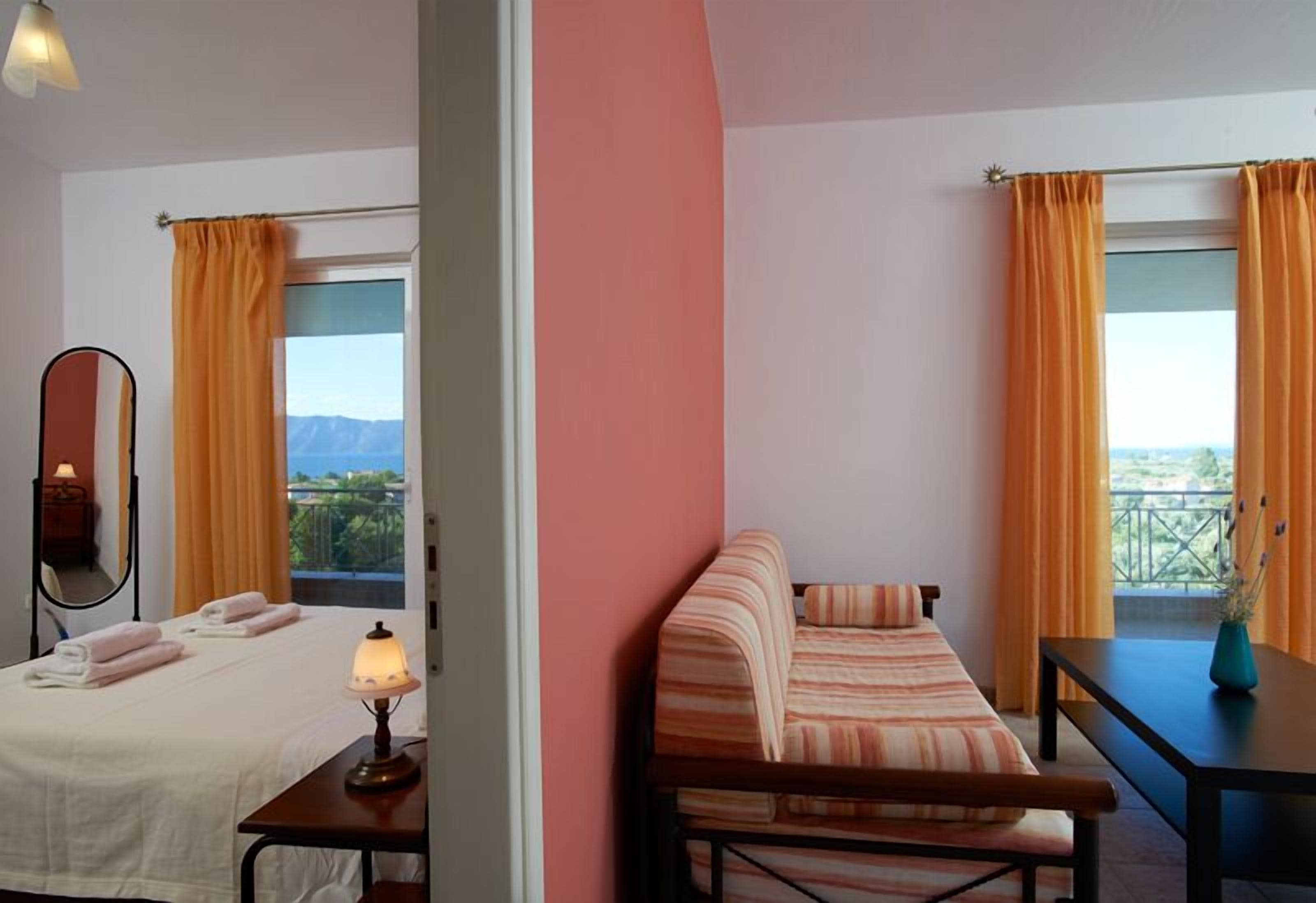 Quarto de hotel com duas camas, um sofá, uma mesa e varanda com cortinas laranja.