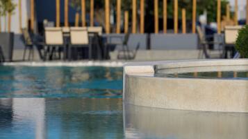 Una piscina con borde de hormigón, rodeada de sillones de descanso y un edificio al fondo.