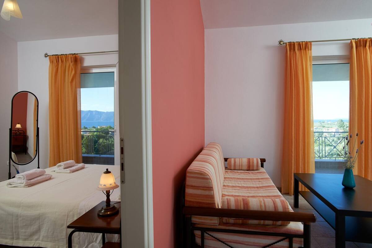 A hotel room with two beds, a sofa, a table, and a balcony with orange curtains.