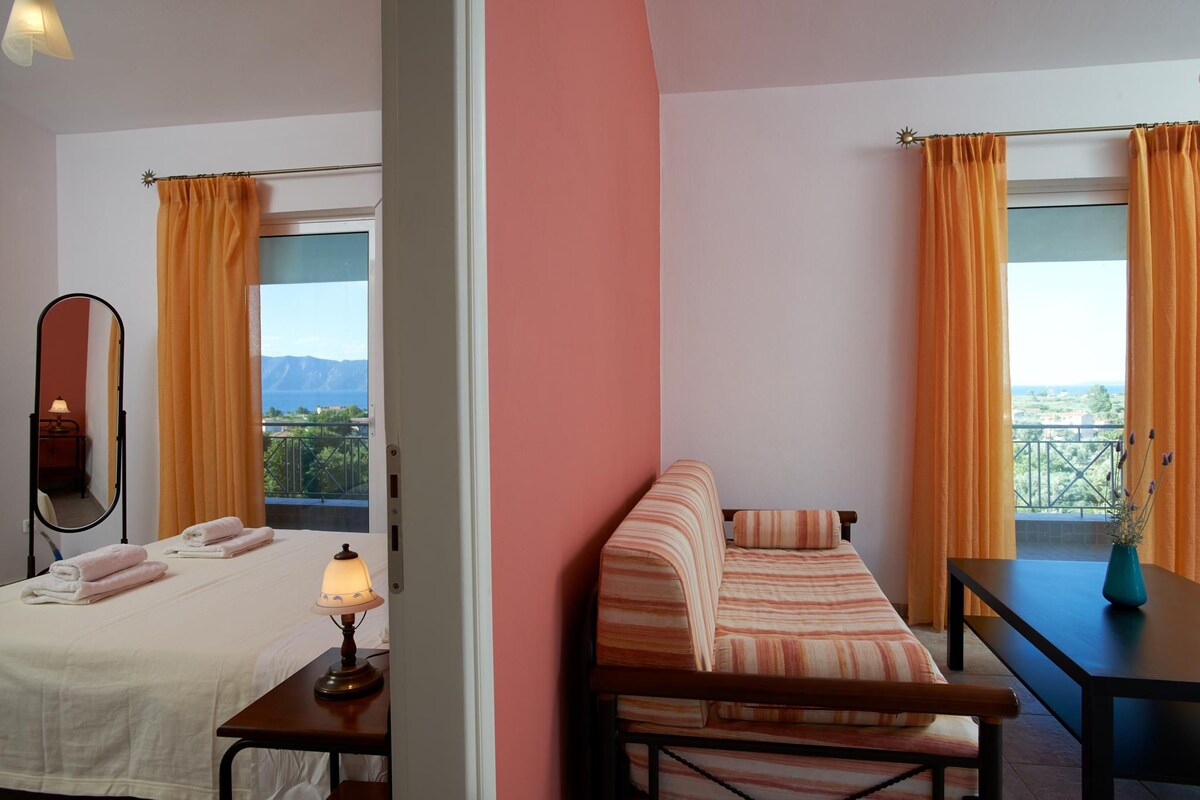 A hotel room with two beds, a sofa, a table, and a balcony with orange curtains.