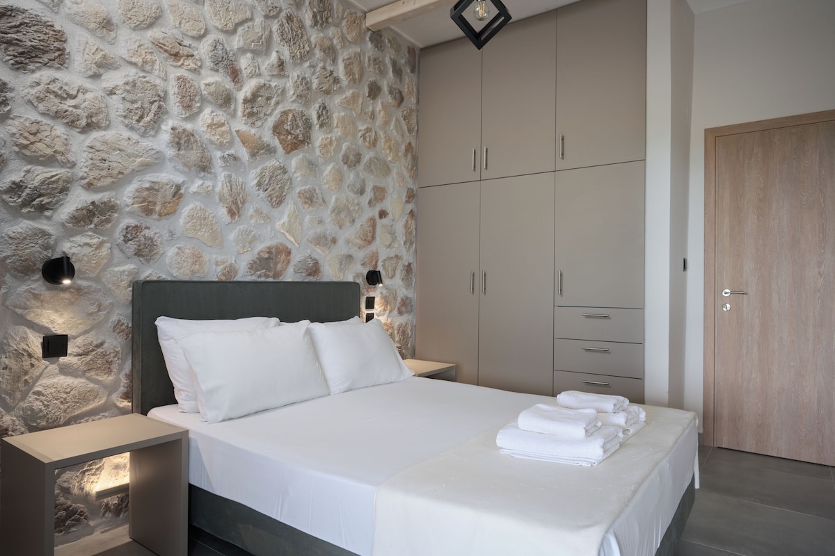 A bedroom with a stone wall, a bed with white linens, a wooden wardrobe, and a nightstand with a lamp.