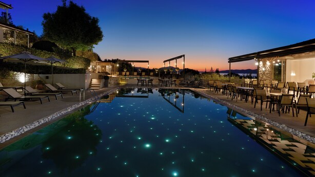Un área de piscina tranquila con sillones y mesas, rodeada de edificios e iluminada por árboles.