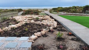 Un jardin aménagé avec une terrasse en bois, une pelouse et un terrain rocailleux qui mène à la plage.