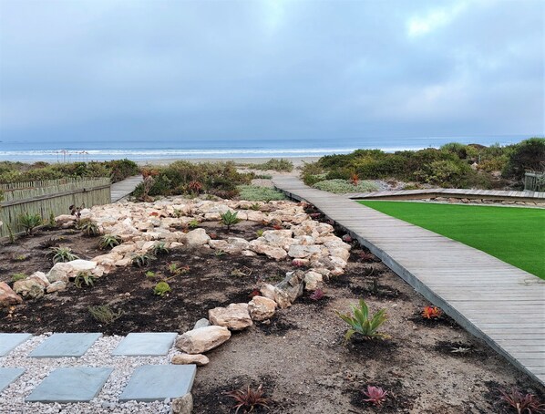 A landscaped garden with a wooden deck, a grassy area, and a rocky terrain leading to the beach.
