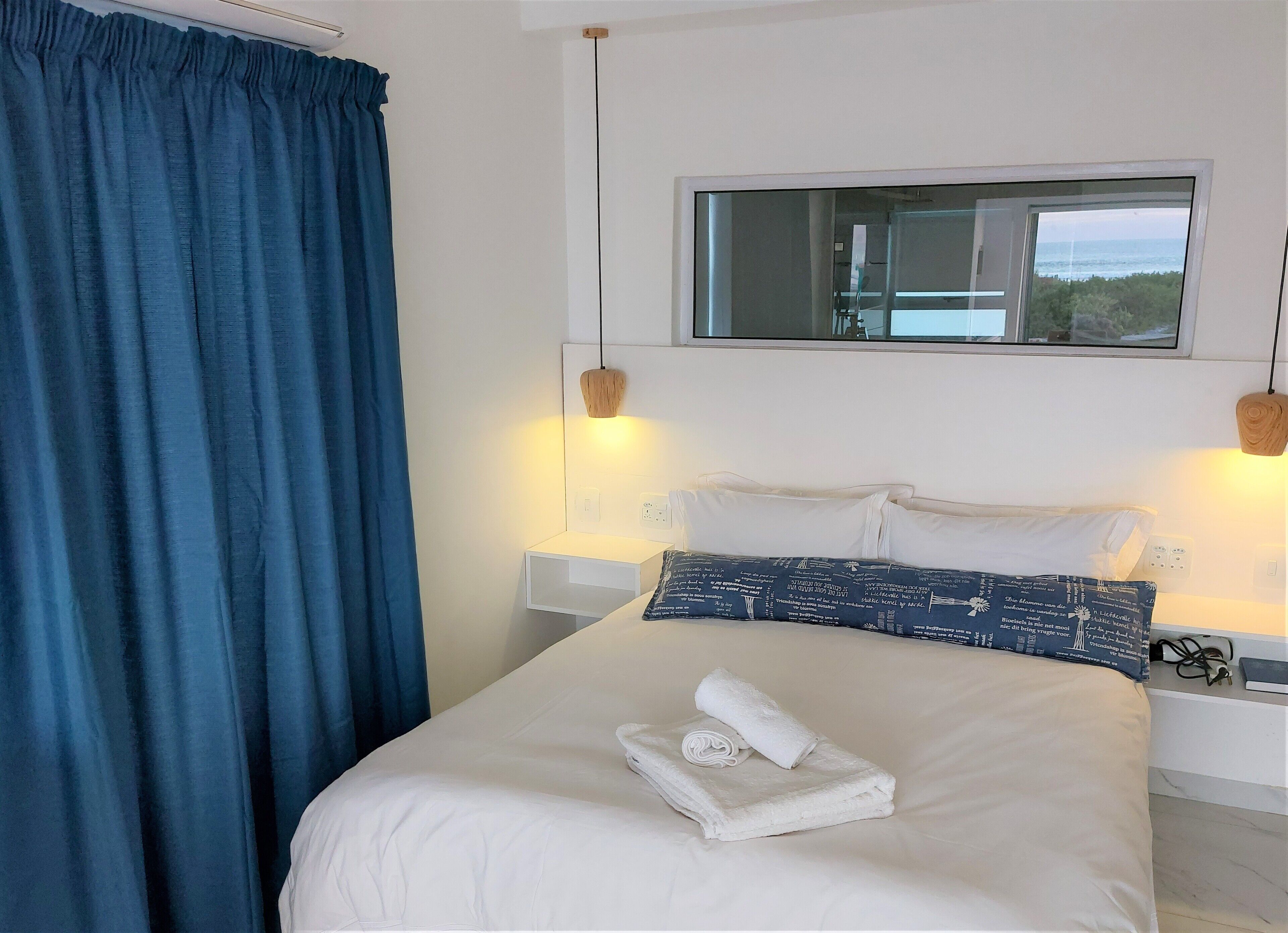 A hotel room with a bed, white bedding, a blue curtain, a bedside table with a lamp, and a window with a view of the sea.