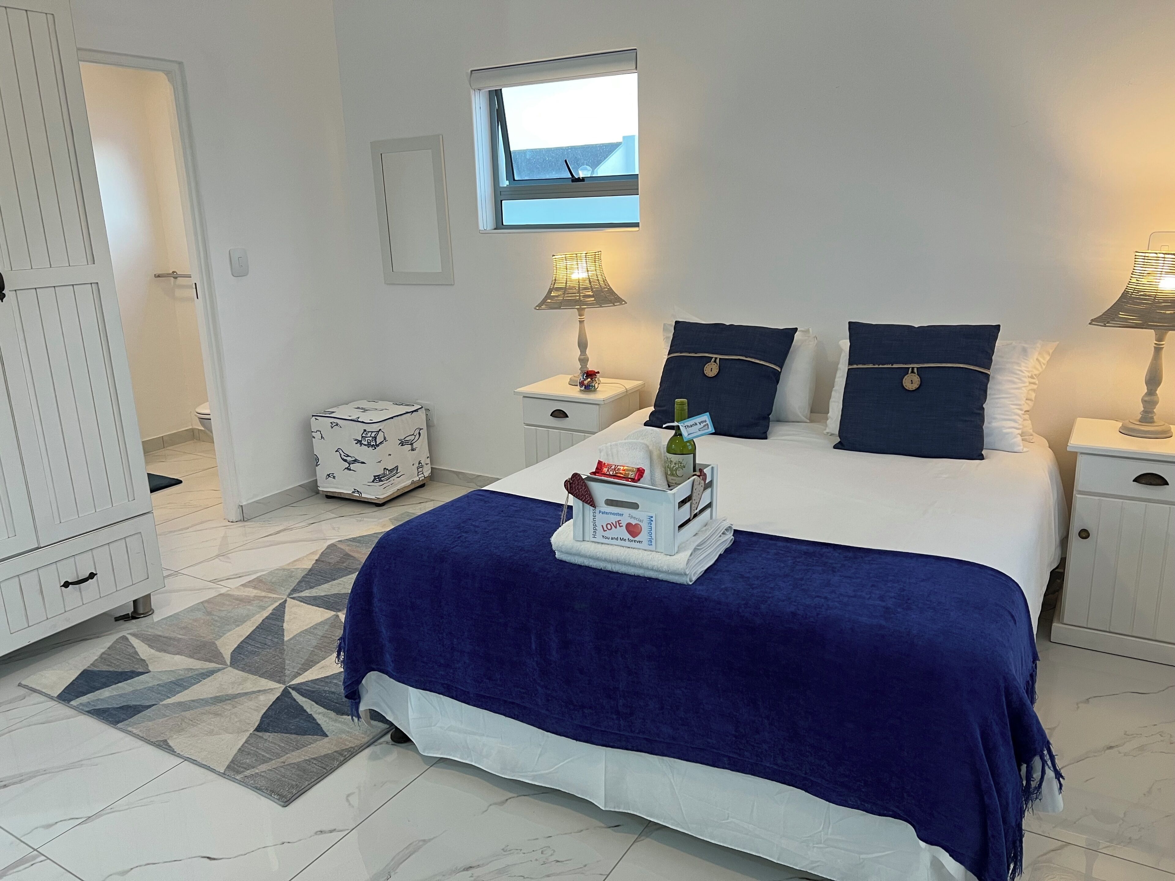 A hotel room with a bed, a nightstand, a bedside lamp, a window, and a small white cabinet.