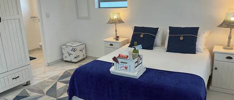 A hotel room with a bed, a nightstand, a bedside lamp, a window, and a small white cabinet.