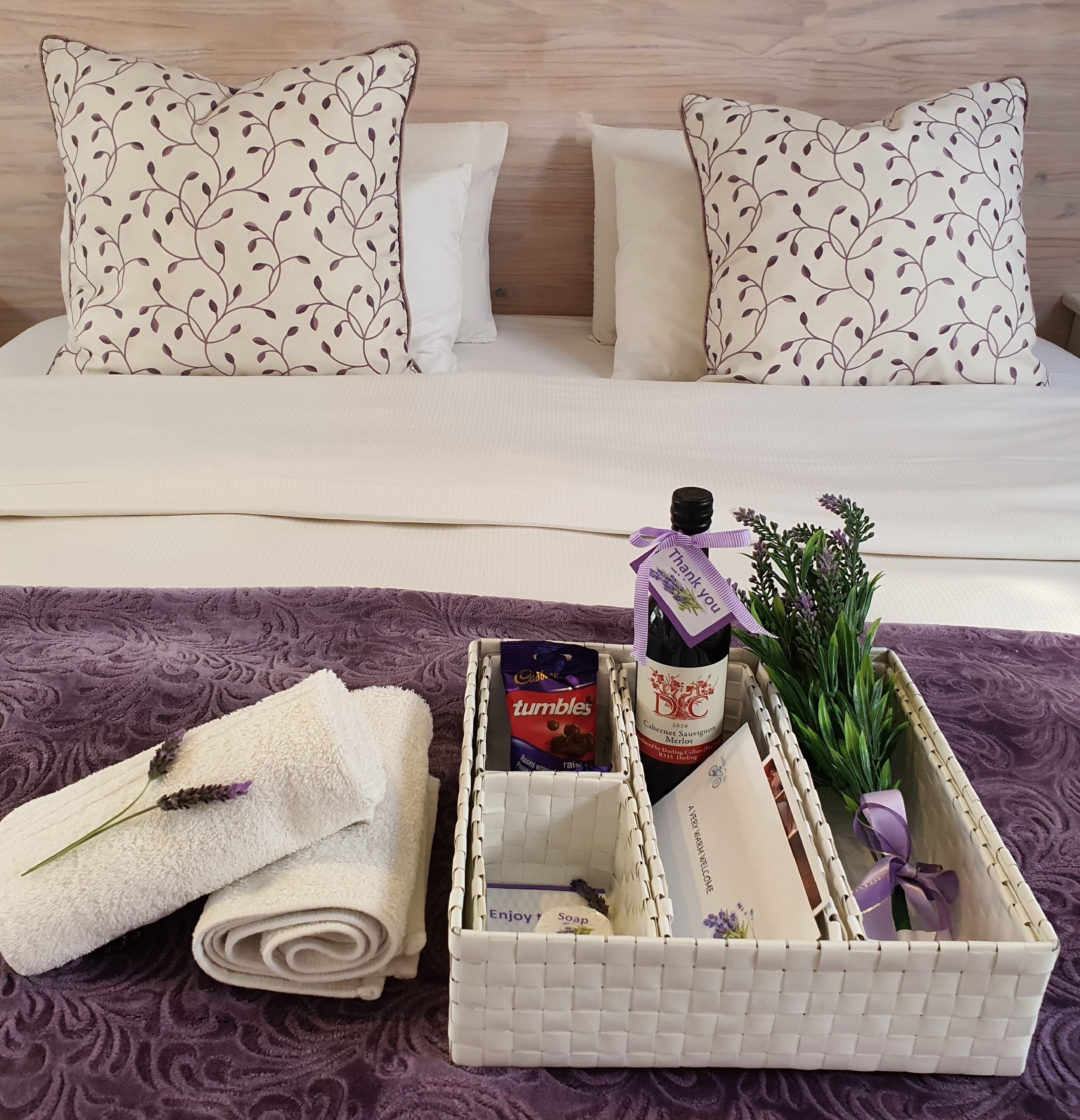 A neatly made bed with patterned pillows, a basket containing a bottle of wine, a chocolate bar, a lavender sprig, and a folded towel on a purple bedspread.