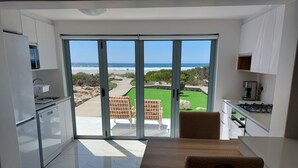Une cuisine moderne avec vue sur l’océan, un coin repas avec des chaises et une terrasse dotée d’un mur en briques et de plantes vertes.