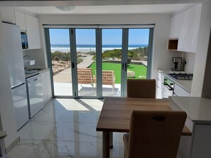 A modern kitchen with a view of the ocean, a dining area with chairs, and a patio with a brick wall and greenery. - Paternoster Accommodation (Paternoster)
