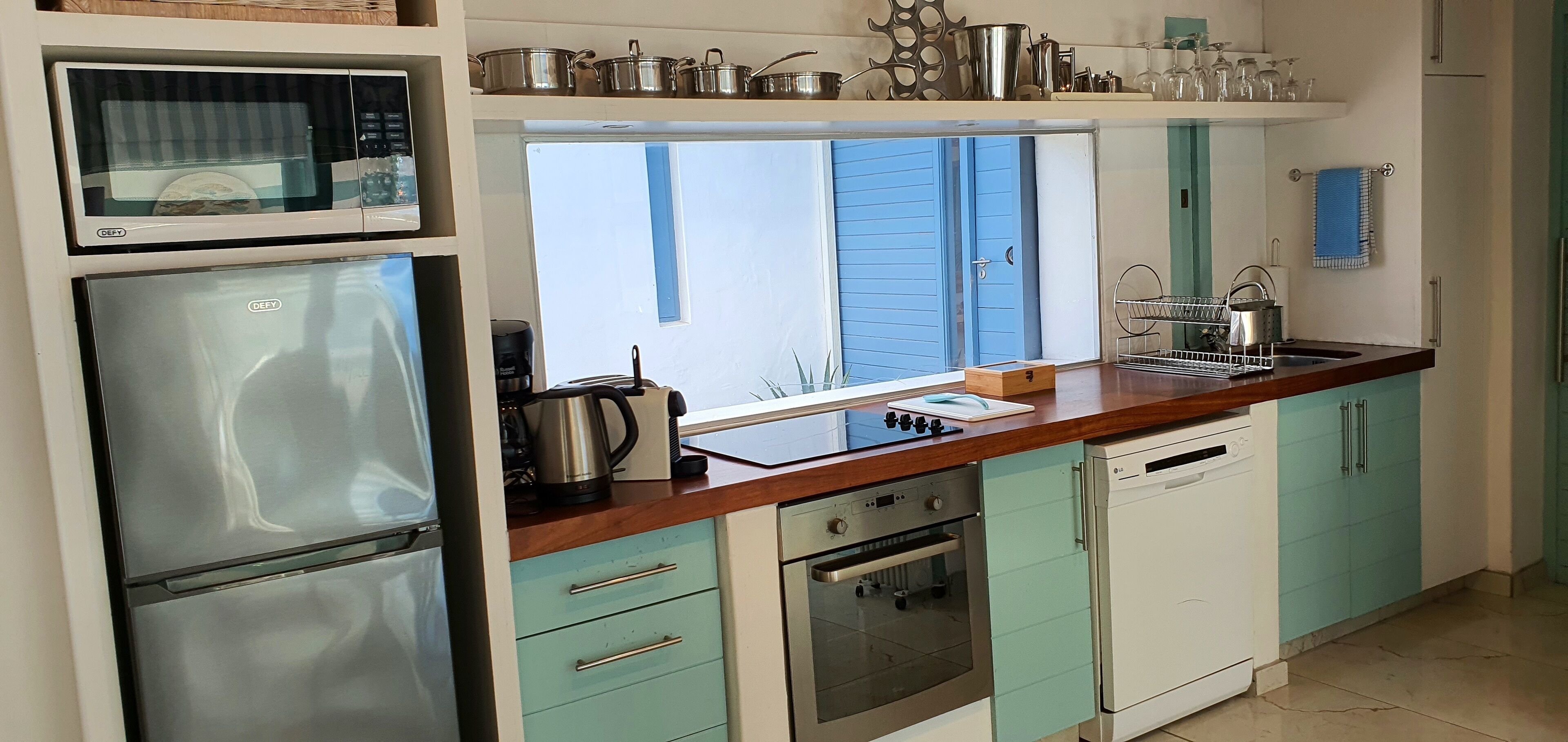 A modern kitchen with stainless steel appliances, teal cabinetry, and a window overlooking a blue door.