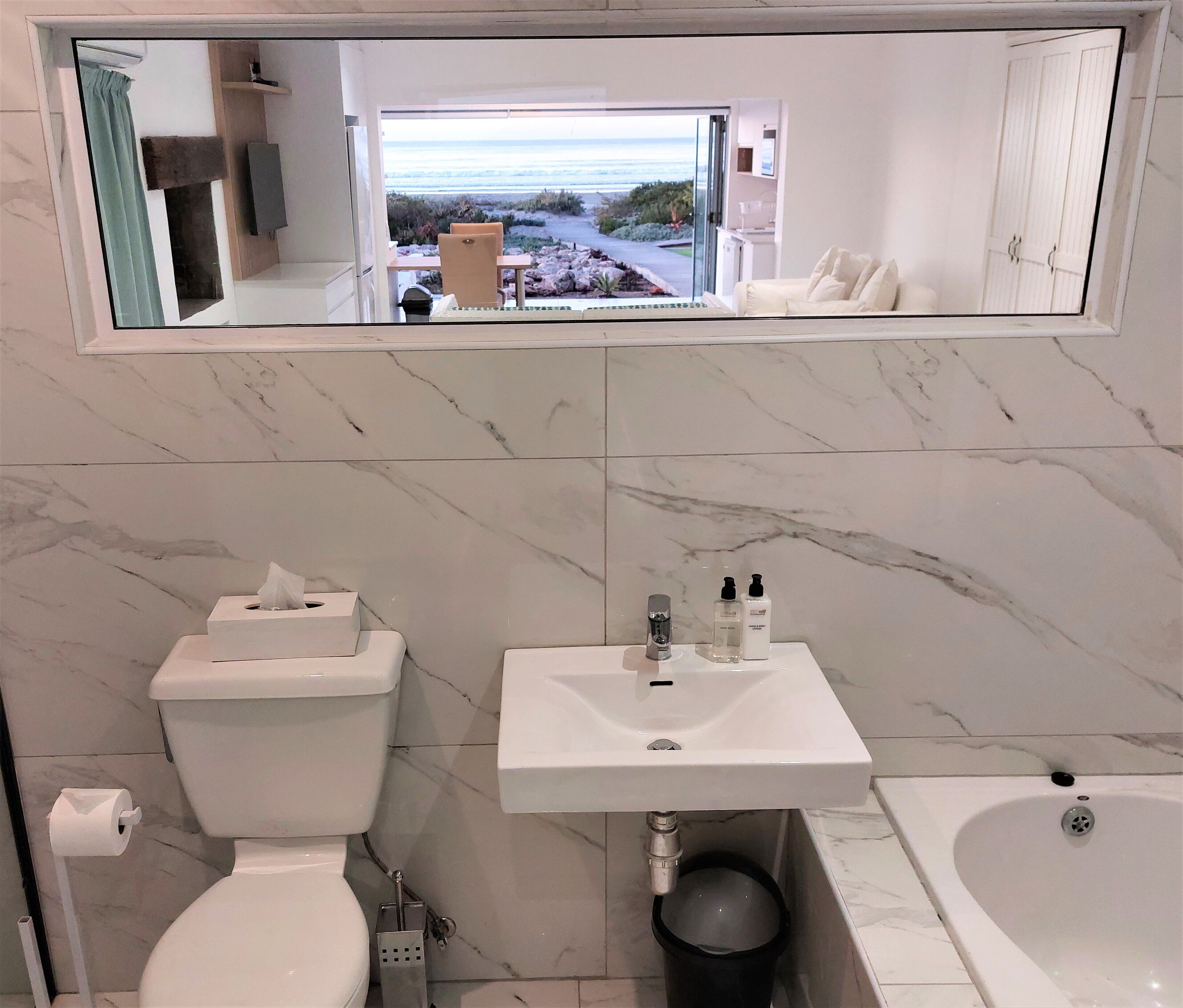 A bathroom with a toilet, sink, and bathtub, a mirror above the sink, and a view of the beach through the window.