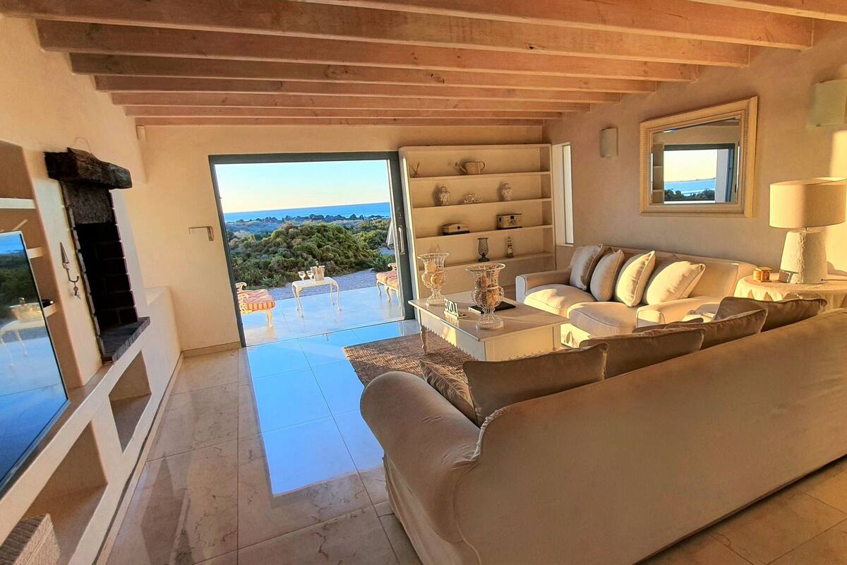A living room with a sofa, a coffee table, and a TV. There is a balcony with a view of the landscape.