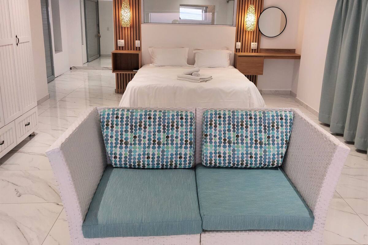 A modern hotel room with a large bed, a sofa with patterned cushions, a wooden nightstand, a mirror, and a window with curtains.