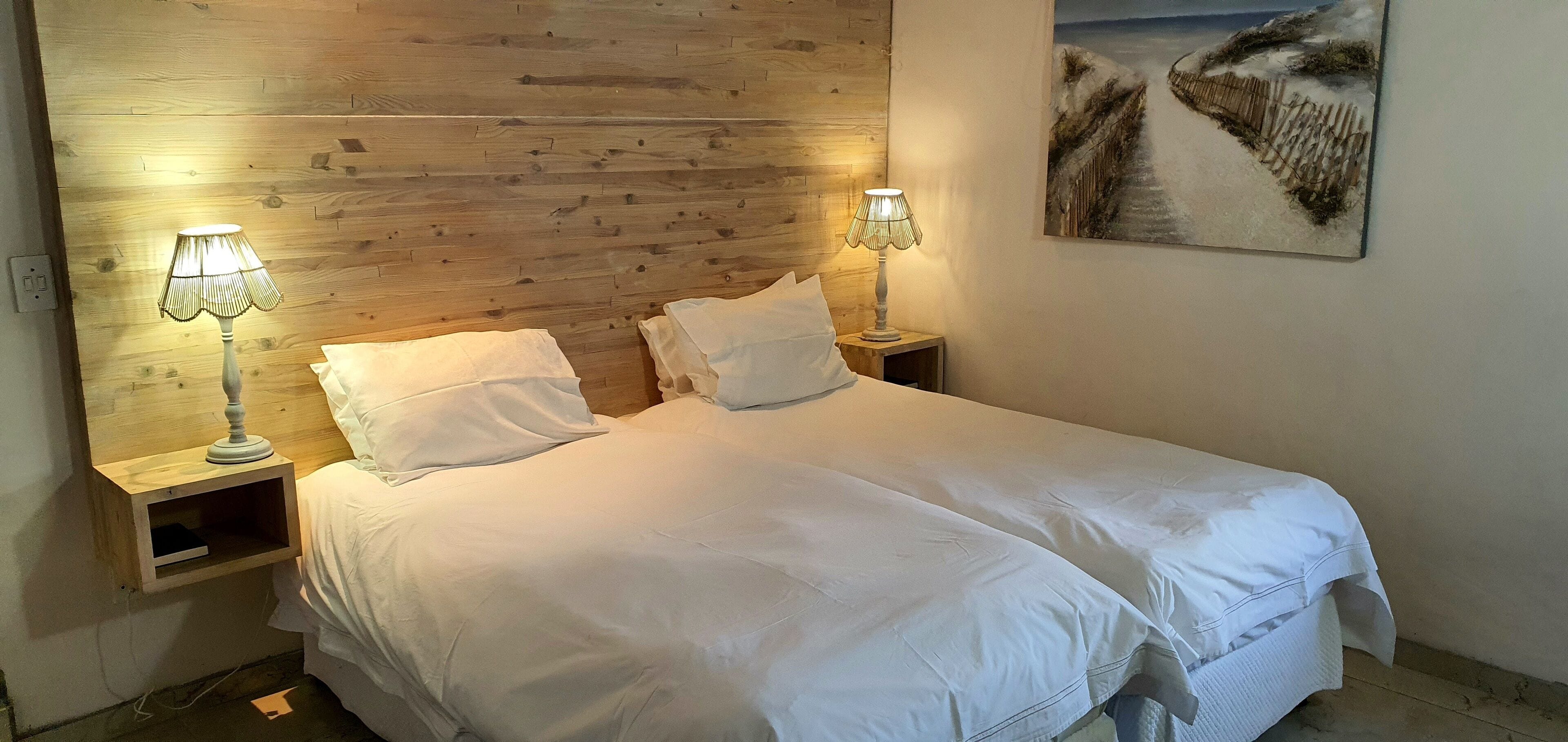A double bed with white linens, two bedside tables with lamps, a wooden headboard, and a painting of a beach scene on the wall.