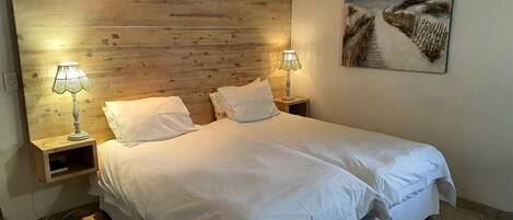 A double bed with white linens, two bedside tables with lamps, a wooden headboard, and a painting of a beach scene on the wall.