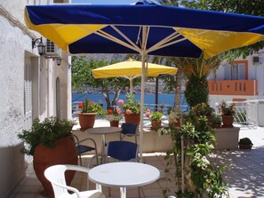 An outdoor seating area with a table and chairs under a blue and yellow umbrella, surrounded by potted plants and overlooking a scenic view. - Valentino Apartments and Studios (Mylopotamos)
