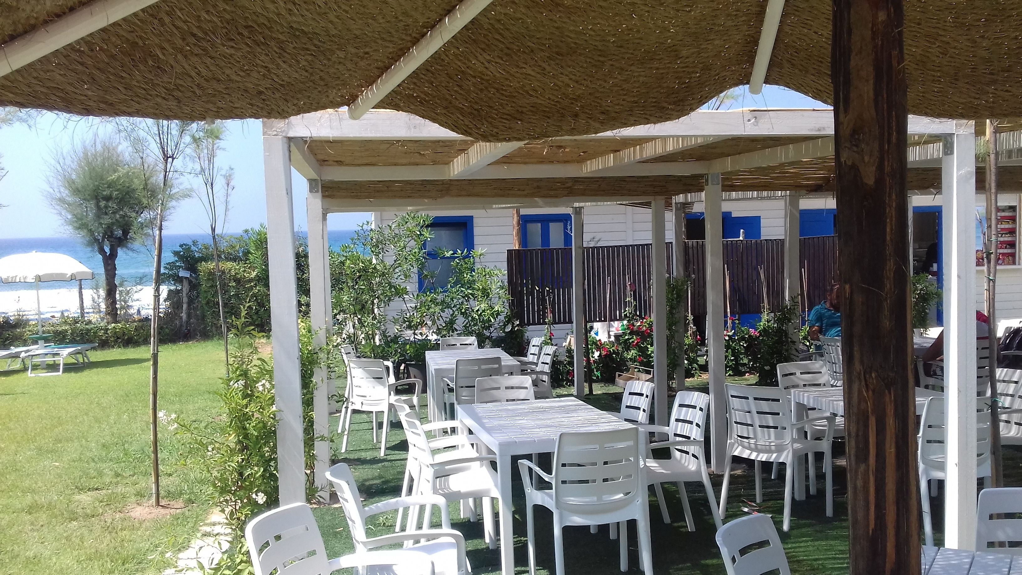 An outdoor patio with white plastic tables and chairs, a thatched roof, and a view of the sea.