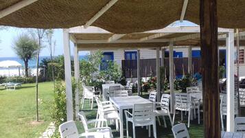 An outdoor patio with white plastic tables and chairs, a thatched roof, and a view of the sea.