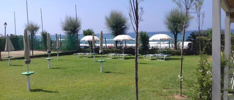 A grassy outdoor area with several white lounge chairs and umbrellas, a fence, and trees.