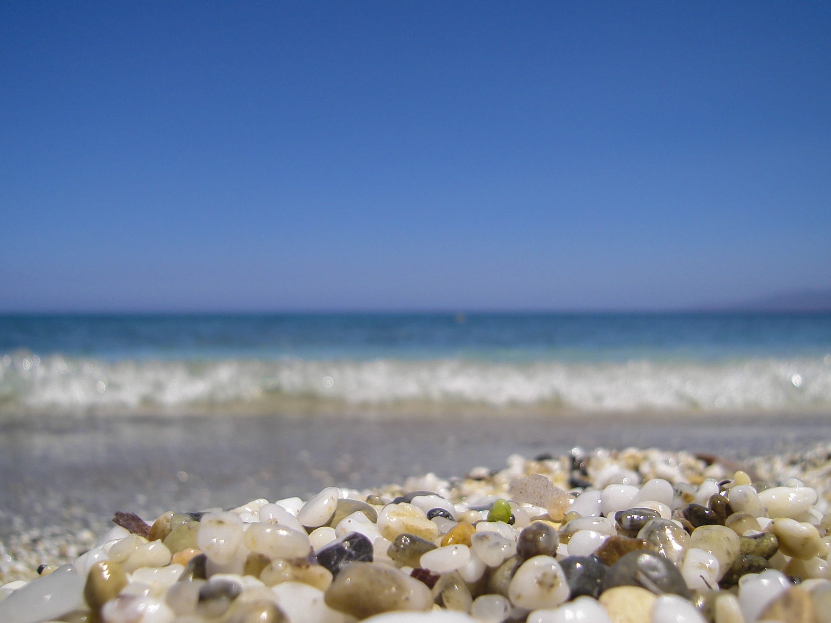A close-up of pebbles on a beach with a clear blue sky and calm sea in the background.