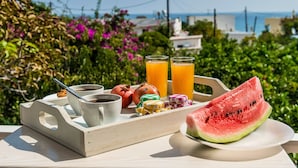 A tray with a cup of coffee, two glasses of orange juice, a pomegranate, colorful candies, and a slice of watermelon on a table with a scenic view of the sea and greenery. - Alpha Stegna Sun (Rhodes)