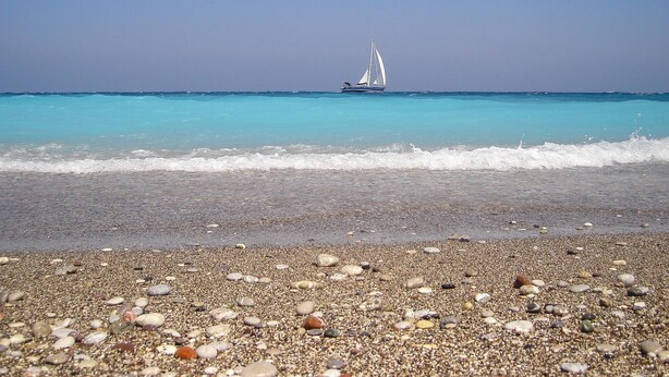 A beach with a sailboat on the water, pebbles on the shore, and clear skies. - Alpha Stegna Sun (Rhodes)