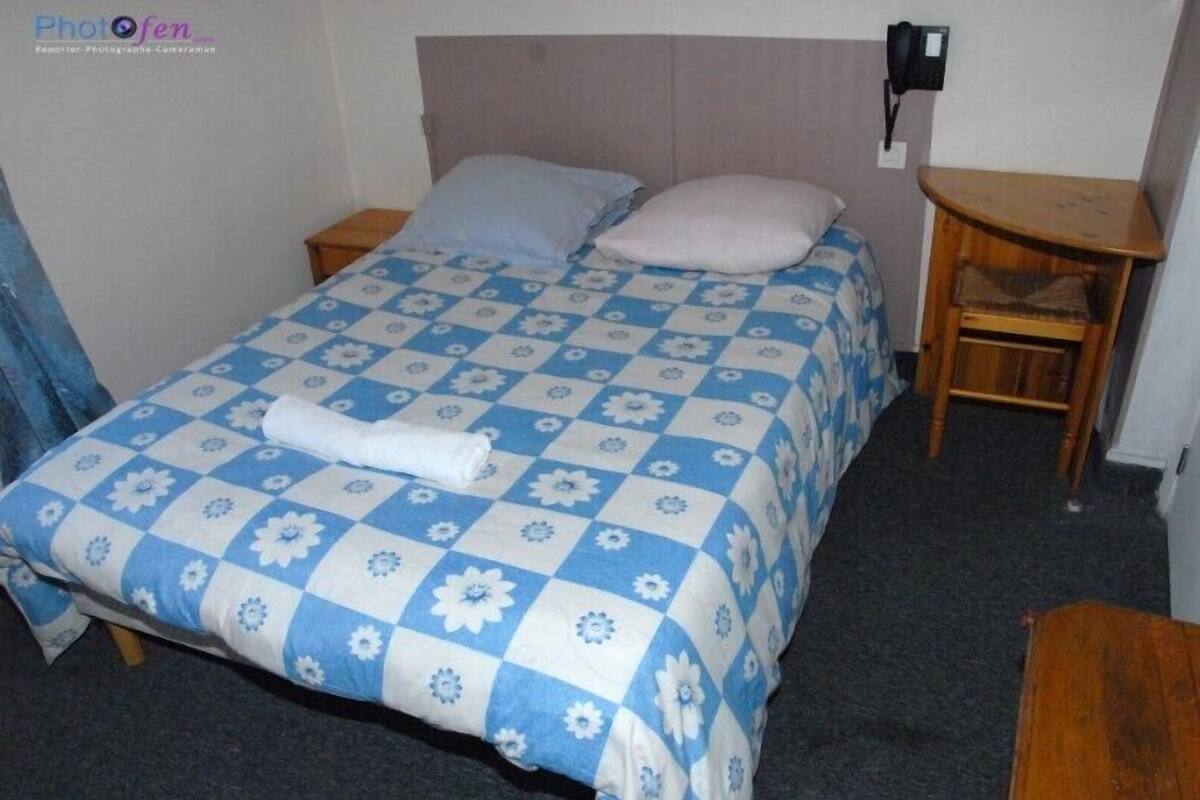 A single bed with a blue and white floral bedspread, two pillows, a wooden nightstand, a small wooden table, and a telephone on the wall.