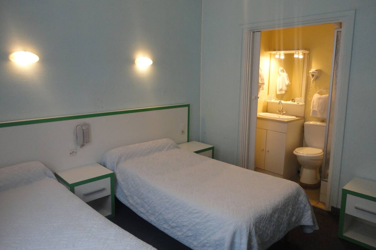 A hotel room with two single beds, a small bedside table with a lamp, and a bathroom visible through an open door.