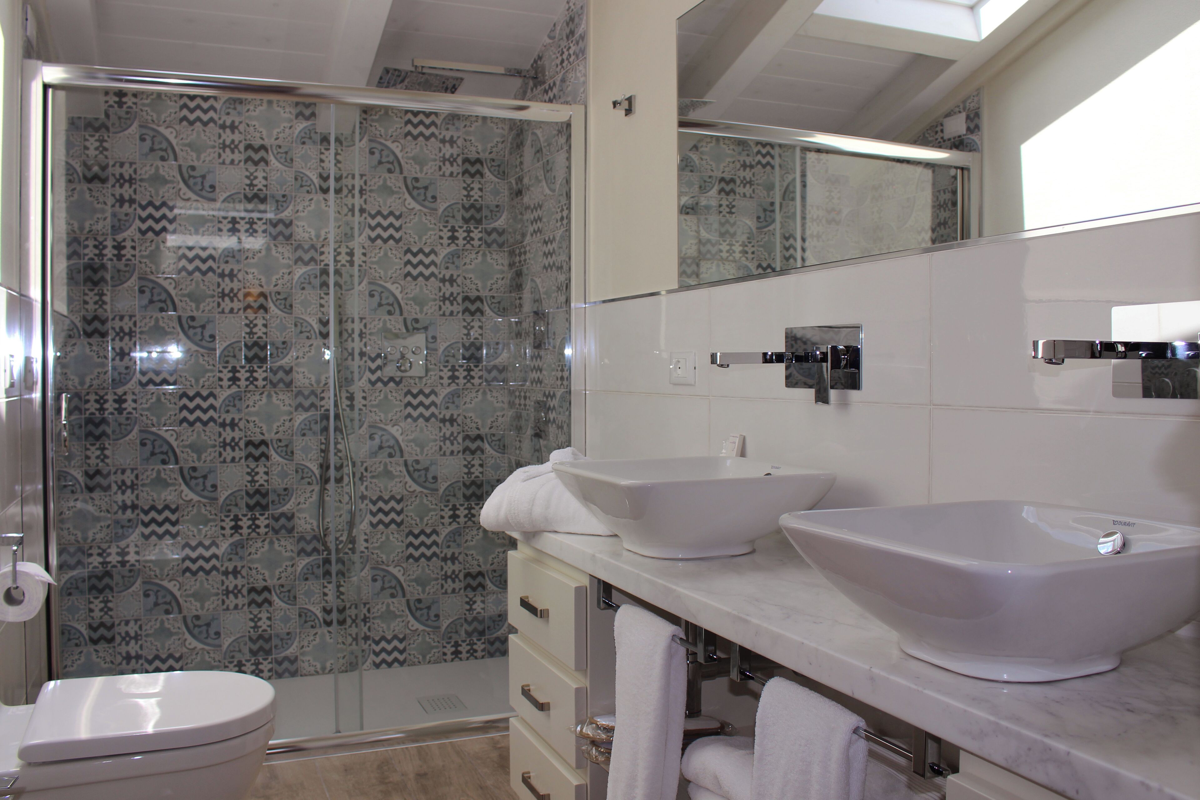 A bathroom with a shower, two sinks, a toilet, and patterned tiles.