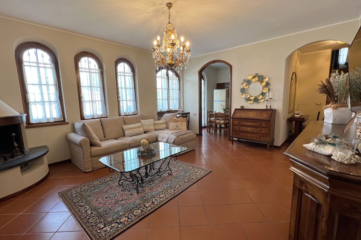 A spacious living room with a large chandelier, a sofa set, a coffee table, and a dining area with a wooden table and chairs.