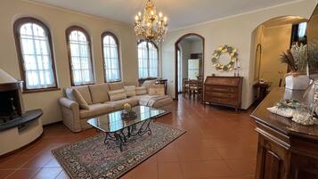 A spacious living room with a large chandelier, a sofa set, a coffee table, and a dining area with a wooden table and chairs.