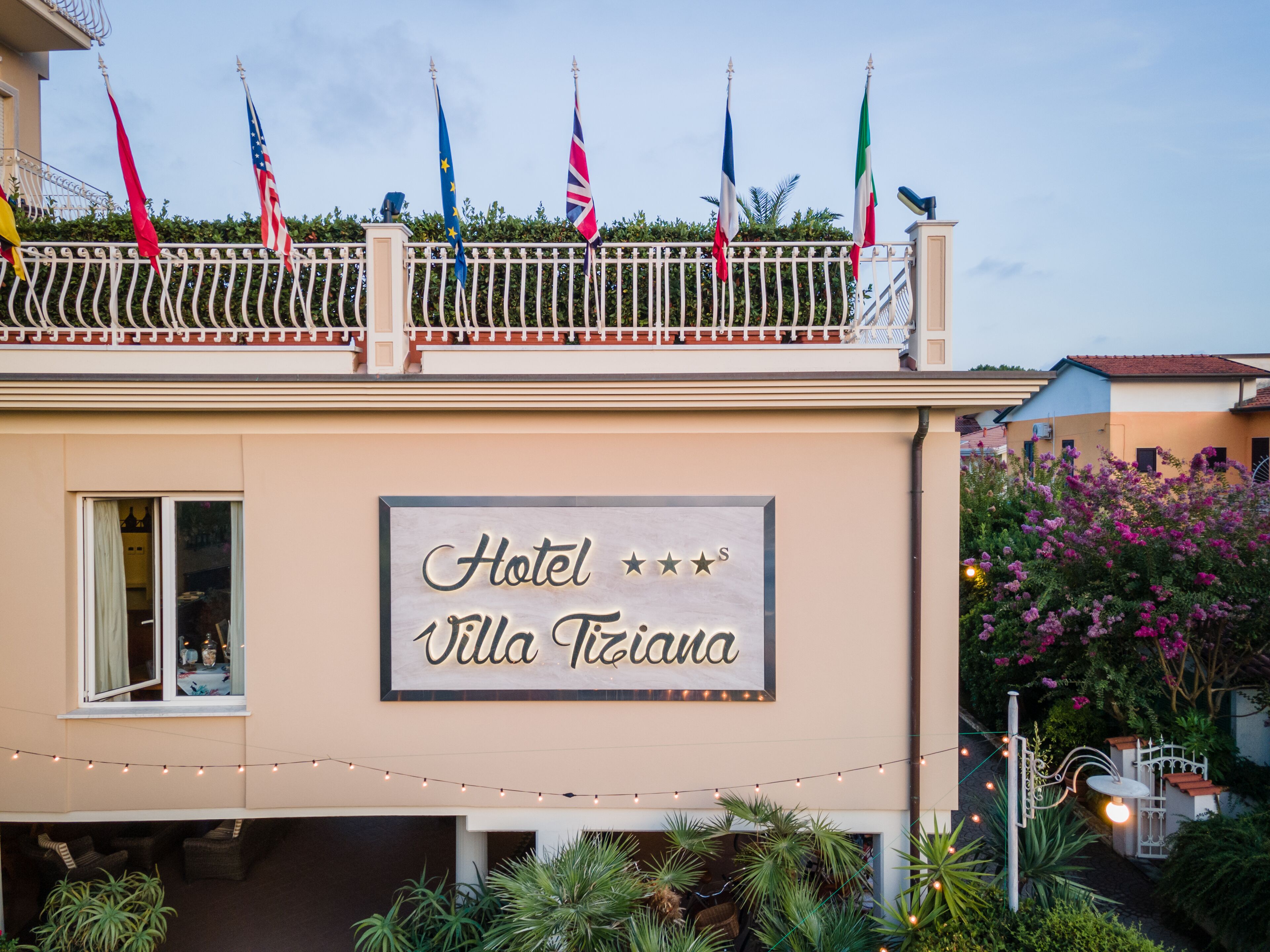 A hotel named Villa Tiziana with a balcony displaying multiple flags, a signboard with the hotel name, and outdoor string lights.