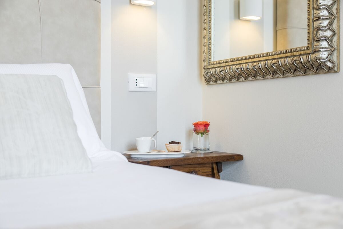 Una habitación de hotel con una cama, una mesita de noche con una taza, un muffin y un florero con una flor.
