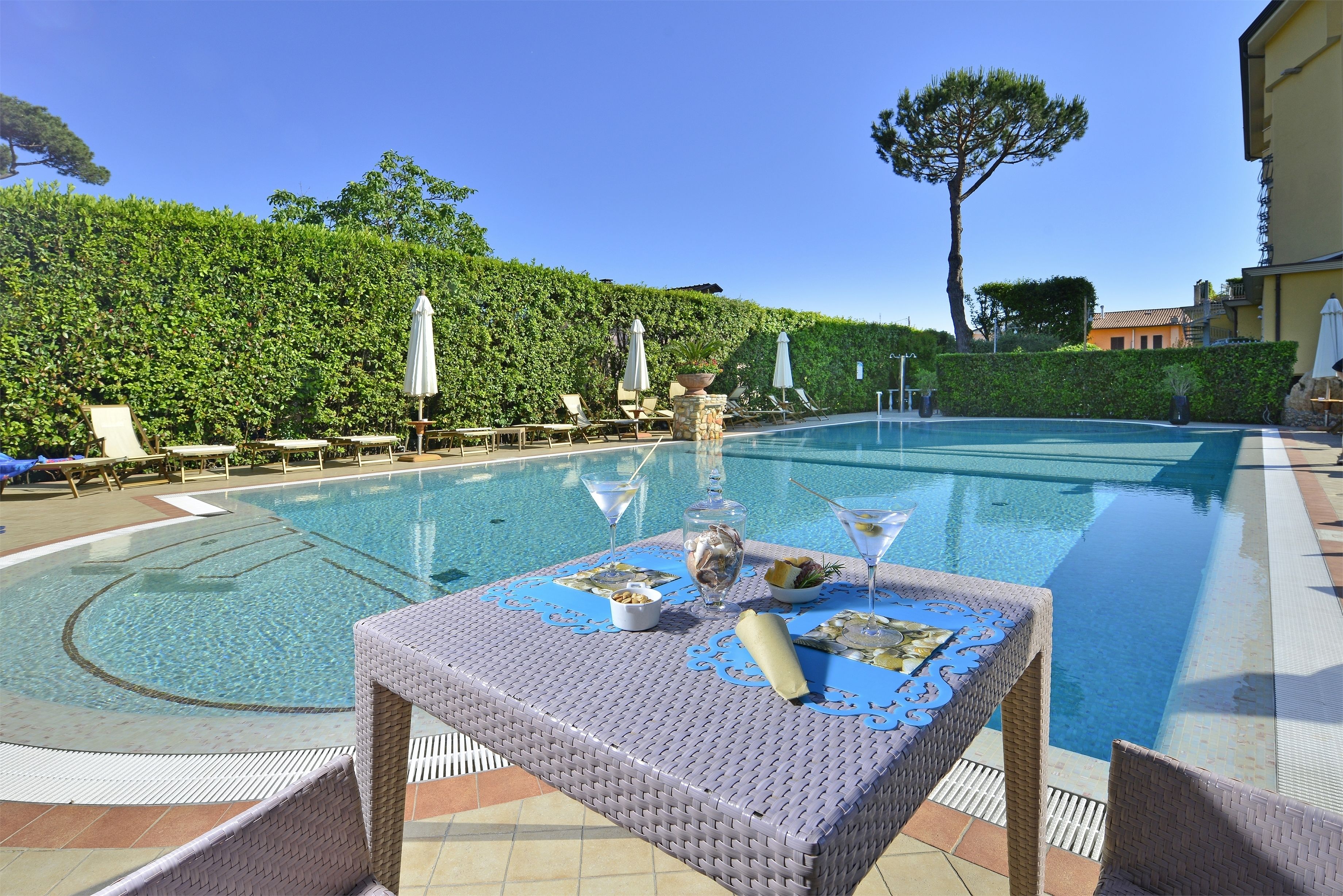 Area piscina con poltrona prendisole, ombrelloni e tavolo con bottiglia e bicchieri.