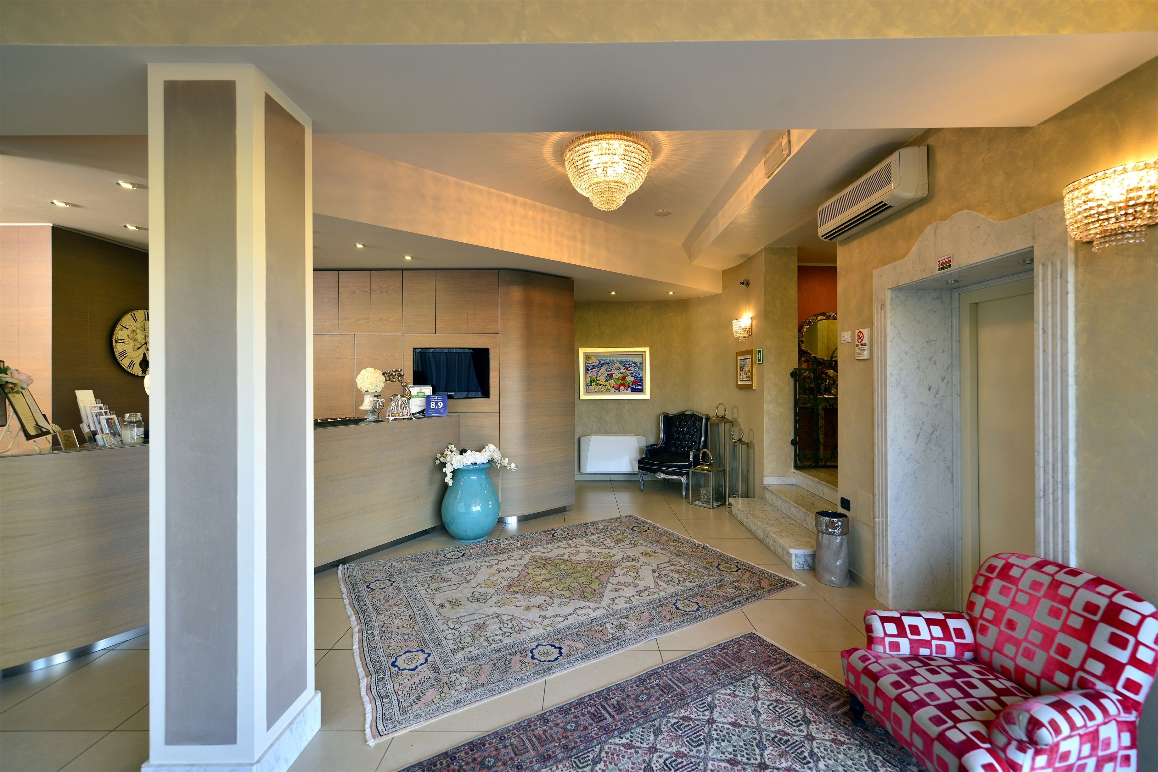 A spacious lobby with a reception desk, a large chandelier, a patterned rug, and a red patterned armchair.