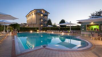 A swimming pool with lounge chairs and umbrellas, a bar area, and a building with a staircase.