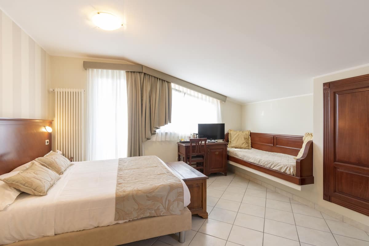 A hotel room with a large bed, a smaller sofa, a desk with a TV, and a window with curtains.
