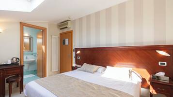 Habitación de hotel con una cama grande, cabecera de madera, escritorio con silla, baño con lavamanos y espejo, y tragaluz.