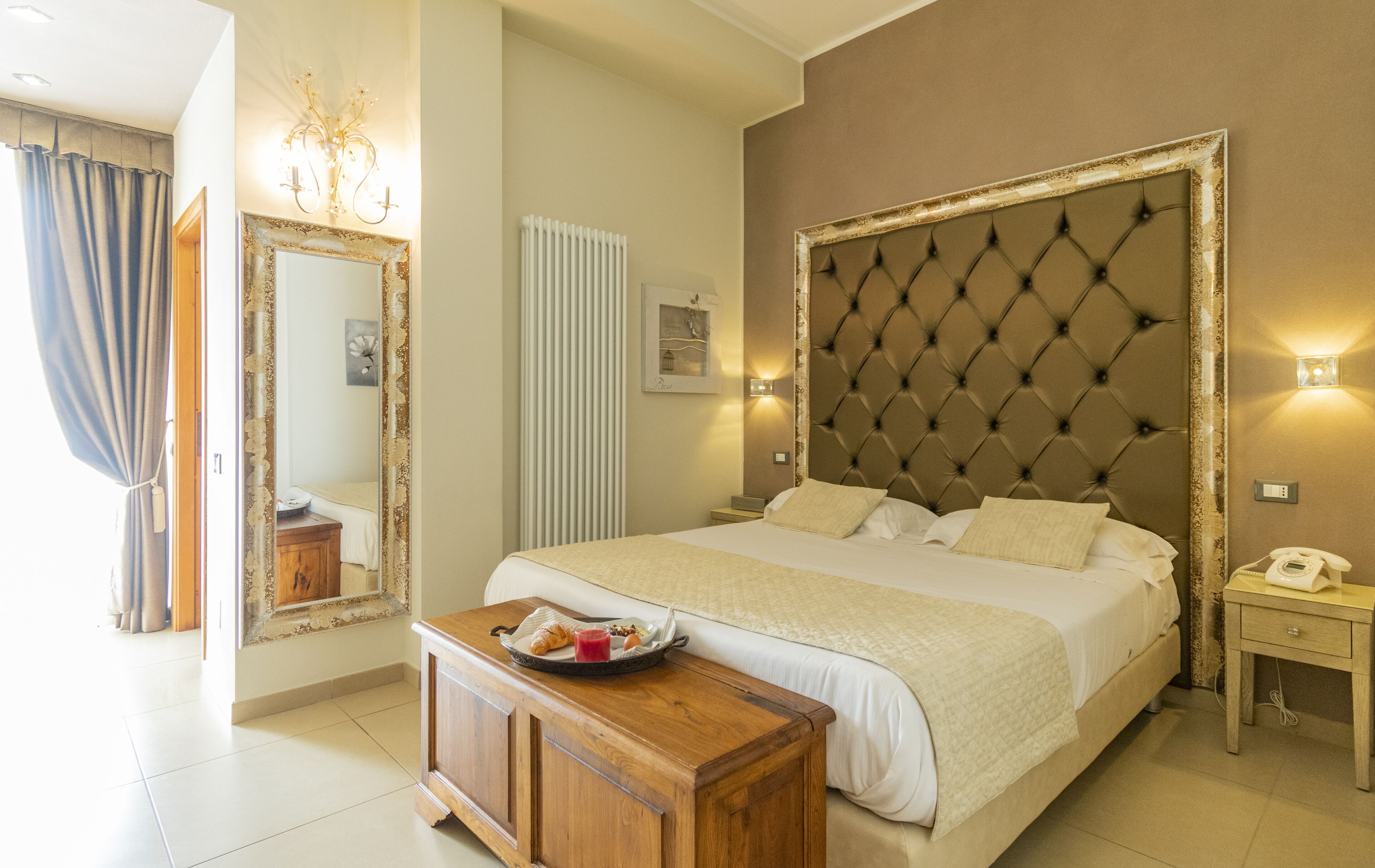 A hotel room with a large bed, a wooden nightstand, a bedside table with a tray of breakfast, a mirror with a decorative frame, and a radiator.