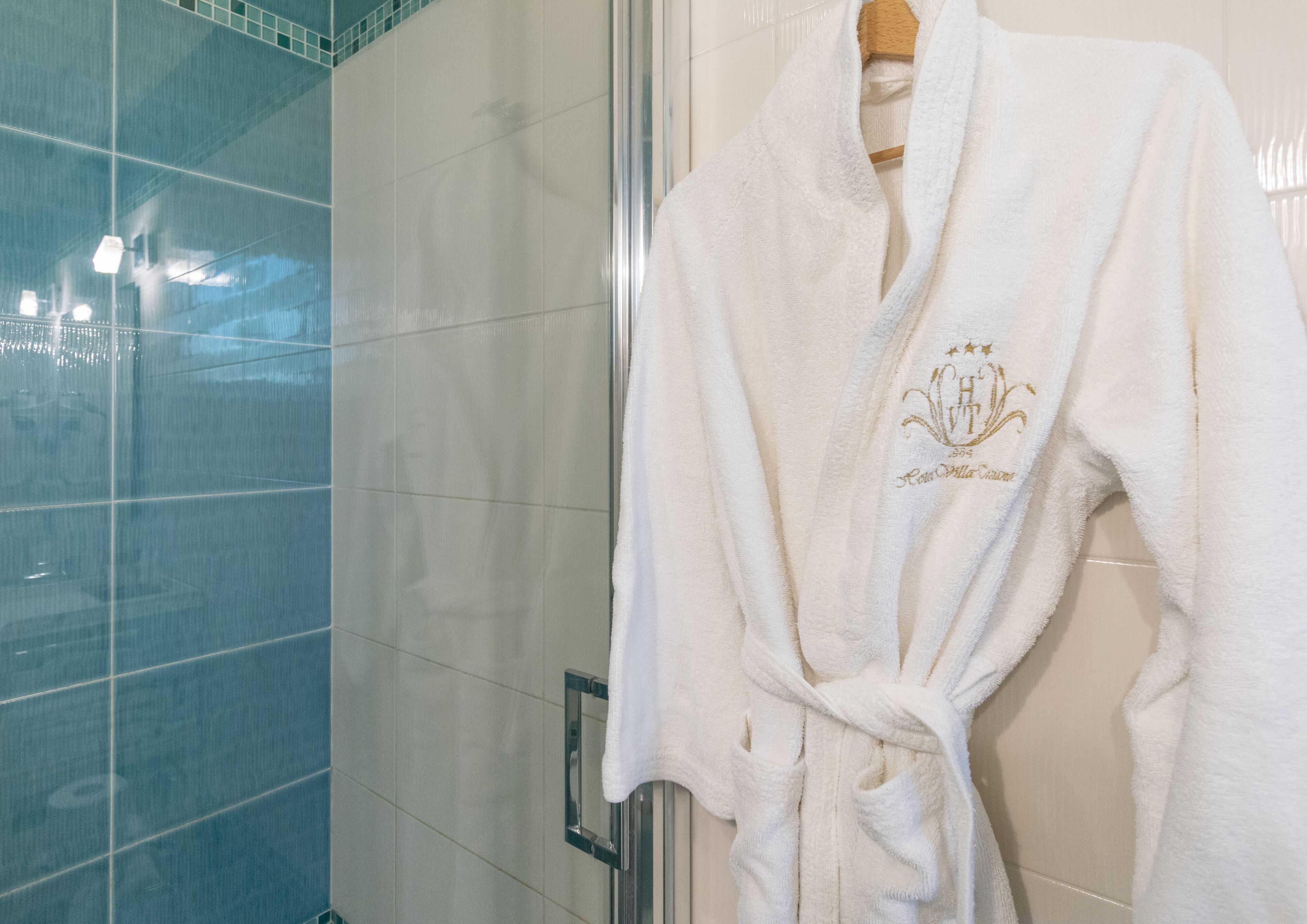 Een witte badjas met een gouden embleem hangt op een rek in een badkamer met een glazen douchecabine.