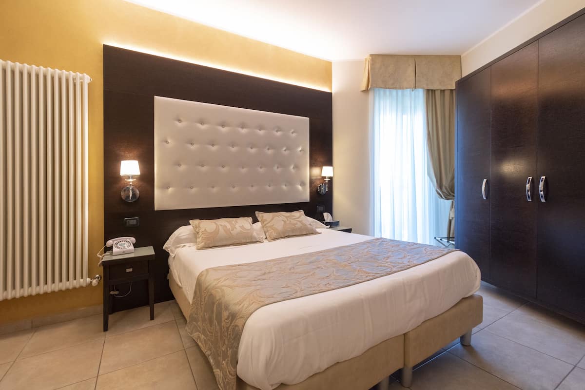 A hotel room with a large bed, a dark wooden wardrobe, a nightstand with a lamp, and a radiator.