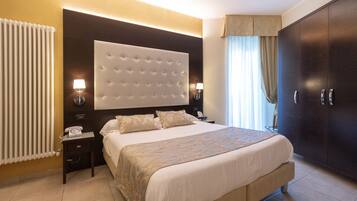 A hotel room with a large bed, a dark wooden wardrobe, a nightstand with a lamp, and a radiator.
