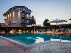 A swimming pool with lounge chairs and umbrellas, a building with a staircase, and a hedge in the background.