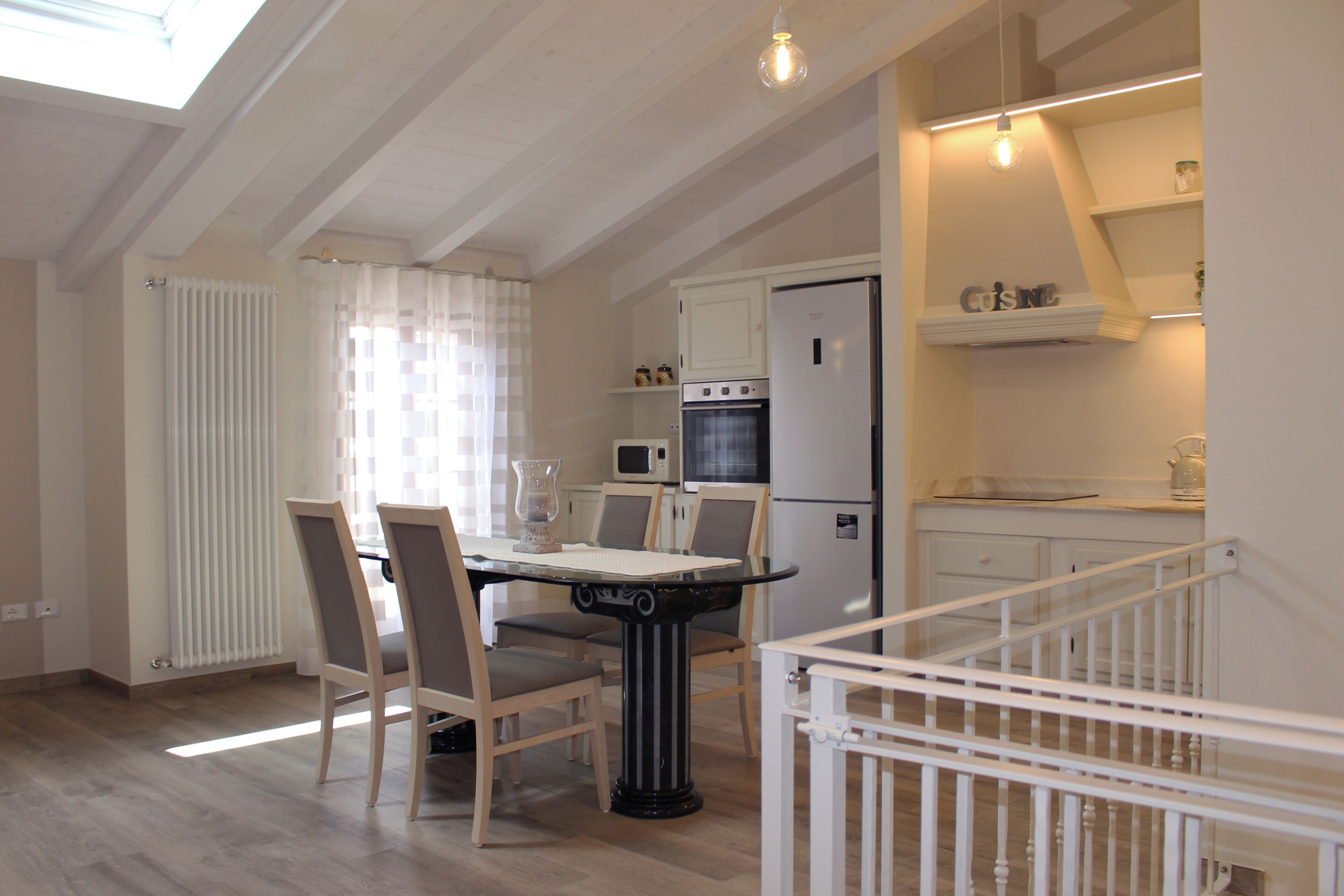 Una cocina moderna con armarios blancos, horno empotrado y microondas. Se aprecia una mesa de comedor con sillas y una barandilla blanca en primer plano.