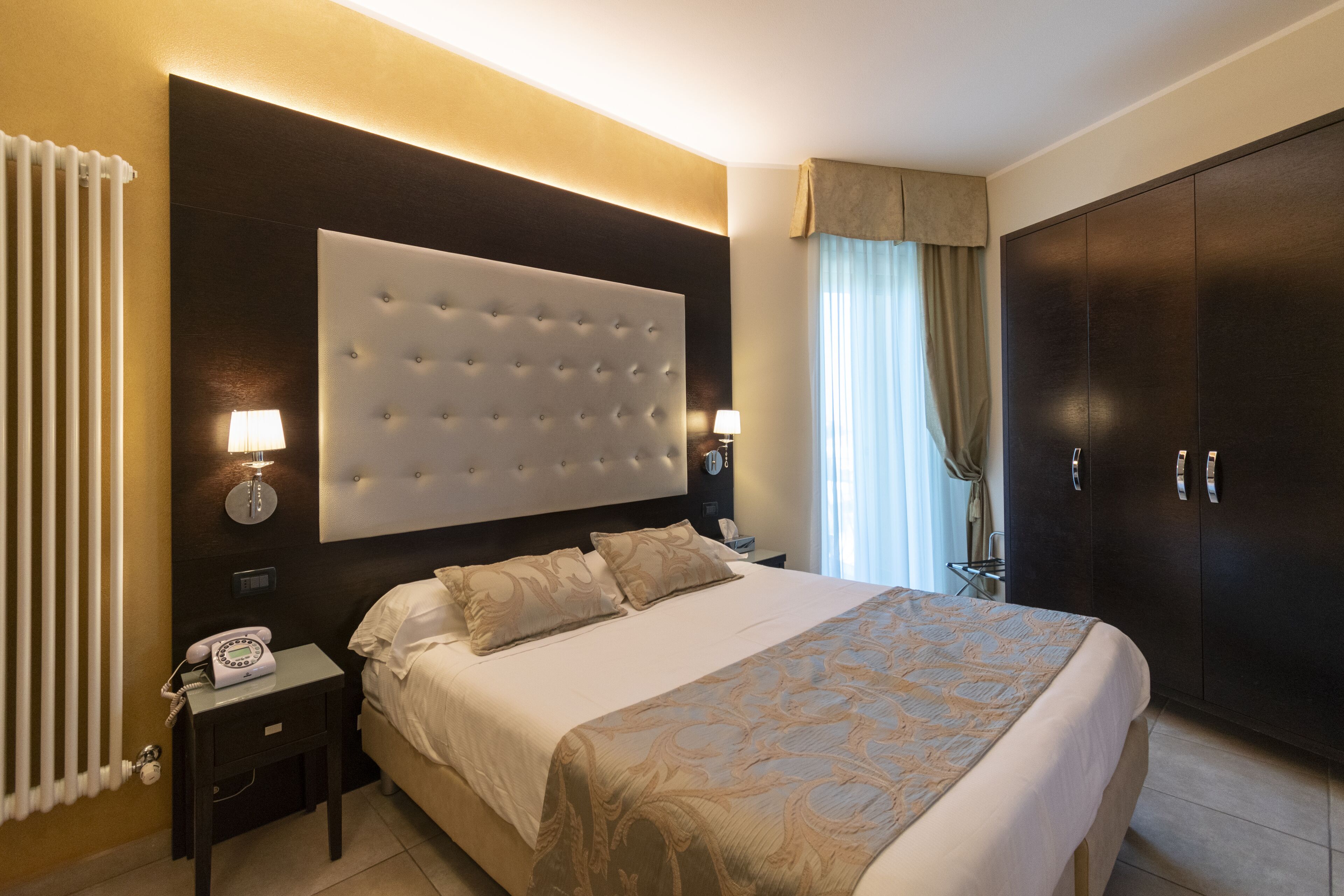 Una habitación de hotel con una cama grande, un cabecero oscuro, una mesita de noche con lámpara, un armario y un radiador.
