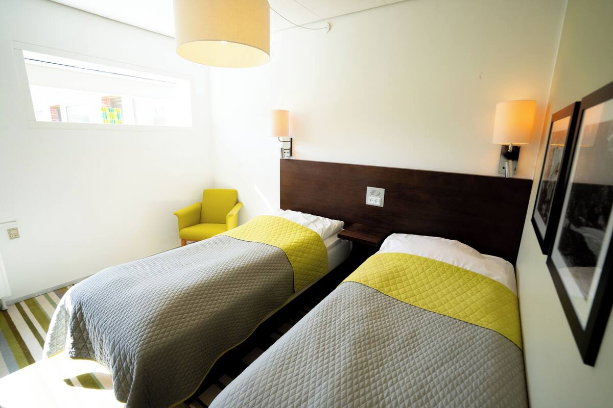 Una habitación de hotel con dos camas, una silla, una ventana y una lámpara fijada a la pared.