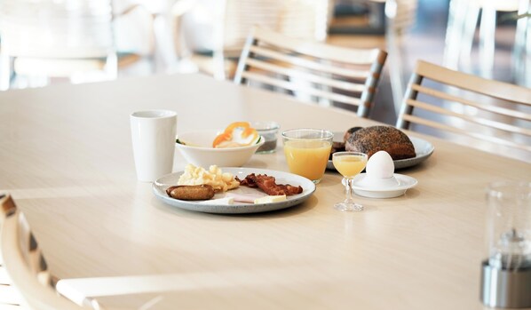 Un desayuno con platos de huevos, tocino y tostadas, un vaso de zumo de naranja y una taza blanca sobre una mesa de madera.