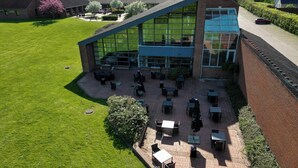Un área de patio al aire libre con mesas y sillas, un edificio de ladrillo con amplias ventanas y un césped verde.