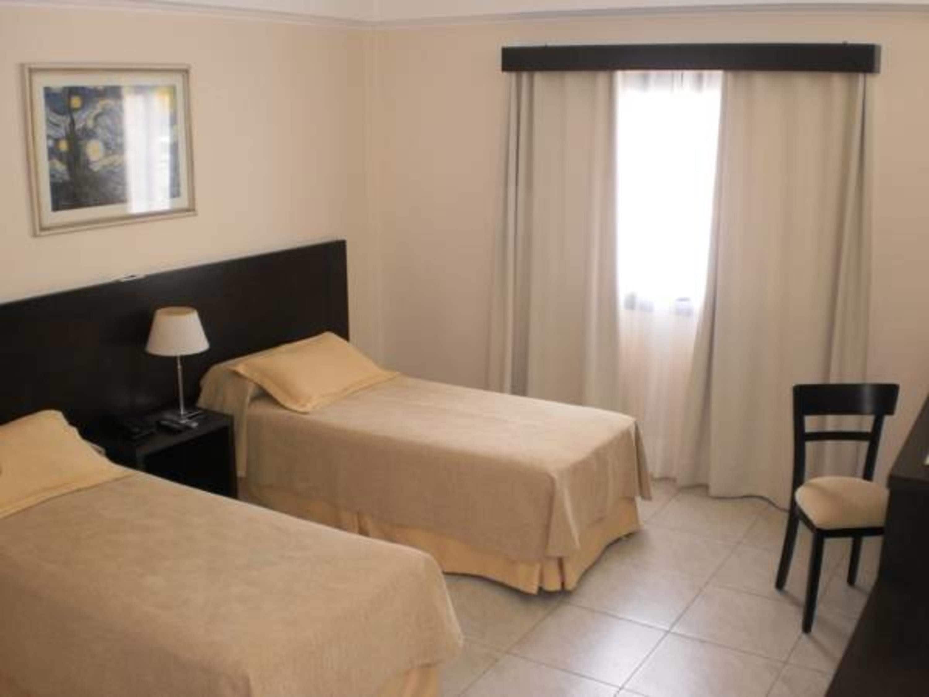 Habitación de hotel con dos camas individuales, un escritorio, una silla y un ventanal con cortinas.